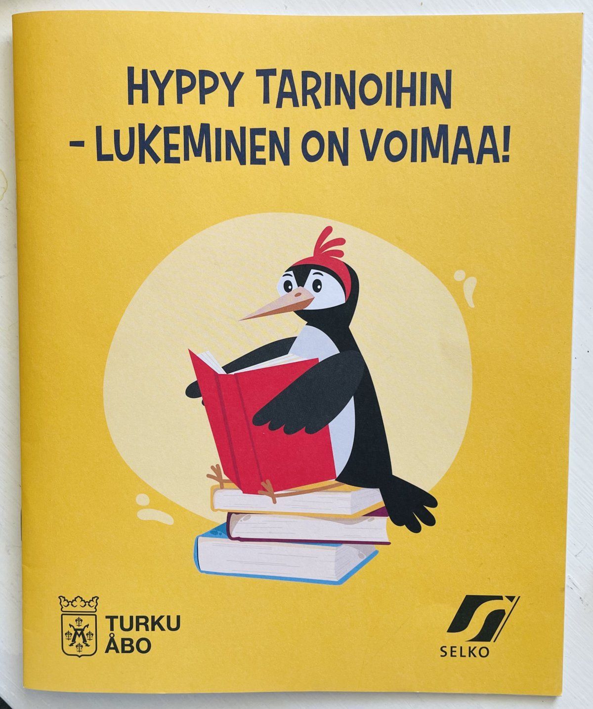 Vihon kansi. Kansi on keltainen ja siinä on lintu lukemassa kirjaa. Kannen tekstissä lukee Hyppy tarinoihin - lukeminen on voimaa!, ja Turun kaupungin logo ja Selkotunnus-logo. 